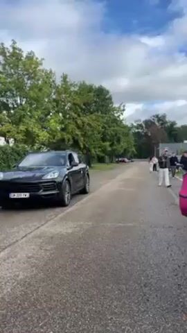 The video begins with a shot of a black SUV parked on the side of a road. There are people walking on the side of the road and trees are lining the street. The camera pans to a group of people standing and talking on the side of the road. The camera then focuses on the side profile of a girl's face. The camera moves again to capture a parking lot full of cars. The camera is being held in someone's hand and we can see their phone.