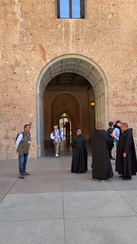 The video shows an outdoor gathering spot at Montserrat Monastery in Spain, with a diverse group of people visible. A man in jeans and a vest stands to the left, while a person in a unique, patterned outfit walks toward the group. Monks dressed in traditional black robes are seen in the center, in conversation. There's a crowd of tourists taking photos and looking around. The monastery's architecture features stone arches, windows with shutters, and balconies. In the distance, a hint of mountainous terrain can be seen, indicating the monastery's location in a mountainous region. The weather appears to be sunny.