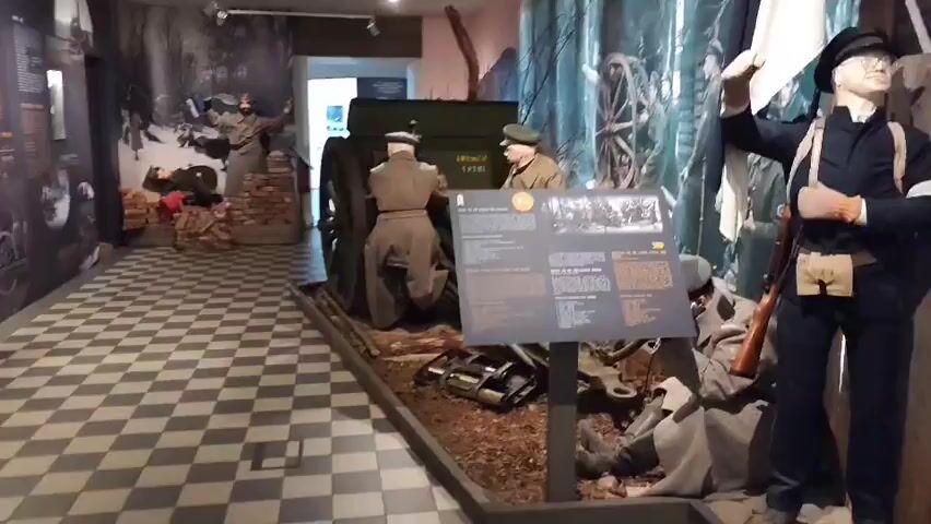The video shows a museum exhibit depicting a World War I scene. In the foreground, a diorama features two mannequins dressed in military uniforms of the era, positioned around a large artillery piece. One mannequin is seated at the breach of the cannon, and the other stands beside it, appearing to operate the mechanism. The diorama is placed on a surface made to resemble ground with logs and debris, and a checkered tiled floor is visible in the background. To the right of the diorama, another mannequin, dressed in a dark uniform with a white band on its arm and carrying a rifle, stands in a more forward-facing pose. Behind the main display, a large mural depicts soldiers and artillery in a wooded setting, adding context to the exhibit. A display board with text and images is positioned in front of the diorama, likely providing information about the exhibit. The lighting suggests an indoor museum environment.