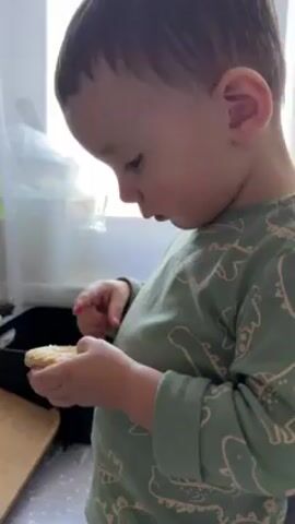 A young child with brown hair, wearing a green long-sleeved shirt with dinosaur patterns, is standing near a window and a kitchen counter. He holds a small, round cookie in his left hand and uses his right index finger to pick at it. The child appears to be inspecting the cookie closely. The background includes a black container and a light-colored surface, possibly a cutting board, on the counter. Sunlight streams in through the window.