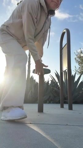 A person wearing a light-colored tracksuit and sunglasses is performing a handstand against a pole outdoors during the day. The person's body is parallel to the ground, supported by their hands on a green object attached to the pole. The background features a bright sky with a tree and large plants, with the sun casting a strong lens flare. The ground is paved.