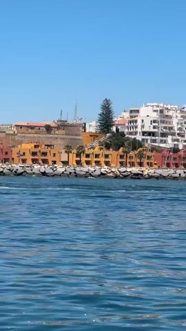The video shows a wide view of a harbor entrance from the perspective of someone on a boat moving through the water. The foreground is dominated by rippling blue water. In the midground, a breakwater made of large stones separates the harbor from the open sea. Behind the breakwater, a line of colorful buildings in shades of orange, yellow, and white are visible. These buildings appear to be apartment complexes or hotels, with a modern architectural style. Among the buildings, tall masts of sailboats are seen, indicating a marina. A few palm trees are interspersed among the buildings. The sky is clear and bright blue. The overall impression is a sunny day in a coastal town with a bustling harbor. The camera is panning slowly to the right, revealing more of the harbor area.