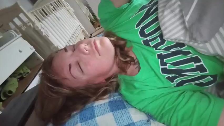 A person with curly brown hair is lying on their side in bed, wearing a bright green t-shirt with dark lettering. Their eyes are closed and they appear to be sleepy or tired, touching their face with their right hand. The background shows a crib with white bars and some stuffed animals visible. The person is lying on a pillow with a blue and white plaid pattern, and a gray and white striped blanket is pulled up over them. The scene is dimly lit, suggesting it is early morning or nighttime.
