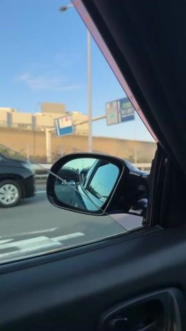 The video shows a car driving on a highway during the day. The view is from inside the car, looking through the windshield and the side mirror. Other cars are visible on the highway, as well as buildings and streetlights along the side of the road. The sky is clear and blue. The dashboard of the car is visible in the foreground.