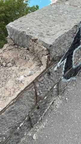 The video opens with a close-up shot of a rugged, weathered stone wall or barrier, likely part of a mountain road or viewpoint. The top surface of the wall shows natural texture and some loose debris. On the vertical face, black spray paint graffiti with white outlines forms stylized, illegible letters or symbols.

As the camera slowly pans upwards and to the right, a breathtaking panoramic view is revealed. The stone wall acts as a foreground element, giving way to a vast landscape below. A deep, winding bay or fjord stretches into the distance, its tranquil blue waters dotted with numerous boats, including several larger vessels that appear to be cruise ships or ferries. Along the shores of the bay, a charming coastal town with densely packed buildings and red roofs is visible, nestled at the base of towering, lush green mountains. These mountains rise steeply from the water, creating a dramatic backdrop under a partly cloudy sky, with patches of blue showing through.

Throughout the short clip, children's voices can be heard speaking an Eastern European language, likely Russian. Phrases such as "А Дуванчики, вы где?" (A Duvanchiki, where are you?) and "А ну-ка повернись" (Come on, turn around) are audible, suggesting a playful interaction among people at the scenic overlook. The time of day appears to be daytime, under overcast but bright conditions. The scene evokes a sense of awe at the natural beauty and the scale of the landscape.