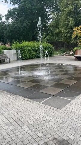 This media file depicts a public park with a splash pad fountain. The fountain is set in a paved area with a checkered pattern of dark and light square tiles. Water jets shoot up from various points on the surface, creating a dynamic display. Several benches are visible in the background, one to the left and another to the right, suggesting a place for relaxation and public use. Lush green trees and bushes form a natural backdrop, with some pink flowers adding a touch of color to the scene. The overall atmosphere appears peaceful and serene, indicating a pleasant day for outdoor activities. The lighting suggests it is daytime, possibly late morning or afternoon, with no strong shadows. The location is identified as Neustadt, Germany. There is no visible text in the media file.