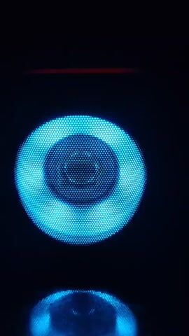 The video shows a close-up of a speaker with a blue light. The light appears to be pulsing or changing in intensity. The speaker is covered by a mesh grille, and the light shines through the holes in the grille. There is a reddish light visible at the top of the frame, above the speaker. The background is dark, suggesting the video was taken in low light conditions.