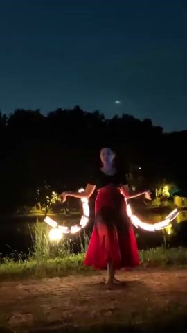 A woman is performing a fire dance at night. She is wearing a black top and a long red skirt. She is holding two flaming hoops, one in each hand, and spinning them. The hoops create trails of light in the dark. She is standing in a grassy area with trees in the background. There is a body of water behind her, and lights can be seen reflecting on the water. The sky is dark blue, and there is a faint moon visible.