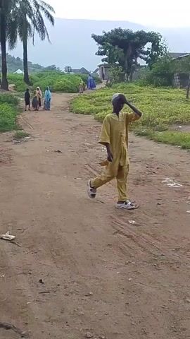 The video shows an outdoor scene on a dirt road. In the foreground, a young boy wearing a yellow outfit and a cap is walking away from the camera, looking towards the right. In the background, a group of people are walking away from the camera on the same dirt road. They appear to be dressed in traditional clothing, with some women wearing headscarves and robes in shades of blue and pink. Palm trees are visible on the left side of the road, and lush green vegetation lines both sides of the path. In the distance, there are some buildings and hills, suggesting a rural or semi-rural environment. The sky appears overcast or hazy. The road has tire tracks and scattered debris.