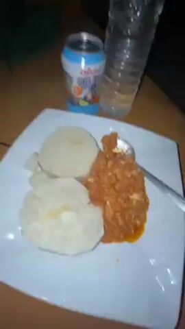 The video shows a plate of food, a can of Calypso drink, and a bottle of water on a wooden surface. The plate contains two large, round, white starchy foods, possibly pounded yam or fufu, accompanied by a reddish-brown stew-like dish served with a spoon. The can of Calypso is light blue and has fruit images on it. The water bottle is clear and partially filled. The lighting is dim, and the camera moves slightly, shifting focus between the food and the drinks.