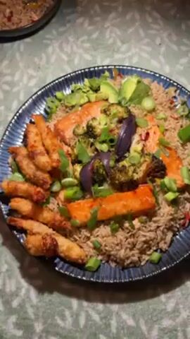 The video shows a plate of food. The plate is blue and has a ridged pattern. The food on the plate consists of brown rice, salmon, fried shrimp, avocado, broccoli, and red onion. The food is garnished with green onions. A bowl of the same rice mixture is visible in the background. The surface that the plate rests on has a green and white floral pattern. A cat meow is audible.