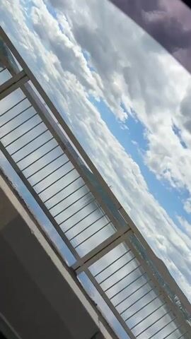 This is a video taken from a moving vehicle, possibly a train or a car, looking out at a sky filled with white clouds against a blue sky. The camera is tilted, giving a diagonal perspective of the scene. In the foreground, there is a structure that appears to be part of the vehicle, with metal beams and rivets visible. There is also a section of translucent or transparent material, possibly a window or a covered walkway, with horizontal bars. Through this structure, the sky and distant land or water are visible. The video is short, lasting only a few seconds, and shows no people or specific actions.