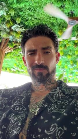 A low-angle shot features a man with a beard, tattoos, and a black paisley shirt, sitting outdoors. He winks and gives a peace sign. The background includes lush green foliage, suggesting a garden or restaurant setting. A light-colored ceiling fan is visible in the upper right corner. The man is wearing two necklaces and looks directly at the camera with a slight smile.