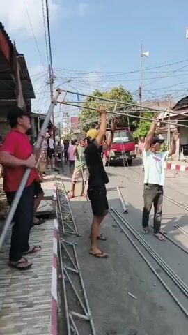 This video shows a group of people assembling a metal structure outdoors, possibly for an event. Several men and one woman are seen lifting and connecting long metal poles, some of which are supported by ladders. The scene appears to be on a street, with houses and a red truck visible in the background. The sky is blue and the lighting suggests it is daytime. The people are dressed casually, and the overall atmosphere seems to be one of coordinated effort and collaboration.