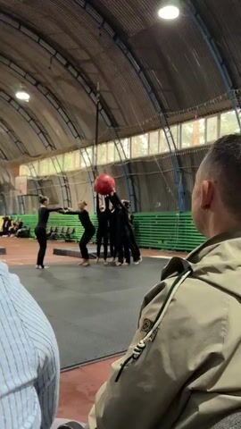 The video captures a gymnastics performance taking place indoors, likely in a gymnasium or sports hall. Several individuals, dressed in matching black athletic attire, are the main subjects. A large, red, inflatable ball is suspended from the ceiling by a rope, and a group of performers are seen pushing it upwards with their hands. Another performer is executing a dramatic, elongated horizontal leap or slide across the floor. Further into the scene, other performers are engaged in acrobatic poses on the floor, demonstrating flexibility and balance. One individual is in a V-shaped pose with their legs extended upwards while lying on their back. In the background, bleachers filled with spectators are visible, suggesting an audience is present for the event. The setting appears to be a large, open space with a high, arched ceiling and visible industrial-style lighting fixtures. Green tiered seating and a basketball hoop are also noticeable, reinforcing the sports venue context. A decorative backdrop featuring a yellow and blue fabric with celestial elements like stars and planets is set up on one side of the performance area, adding a thematic touch to the routine. The overall atmosphere appears to be one of focused athletic performance and artistic expression. There is no discernible text visible in the video.