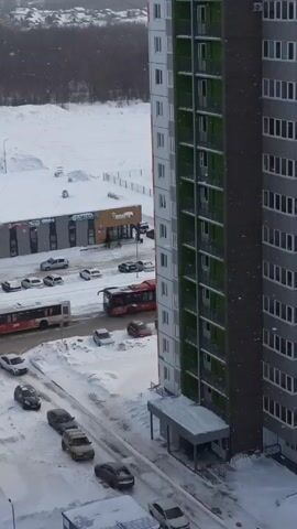The video shows a snowy urban landscape from an elevated perspective, possibly from within a building.  A tall, modern apartment building stands prominently on the right side of the frame, featuring a combination of white, grey, and green accents. The foreground shows a snow-covered street with several parked cars and a few moving vehicles, including a bus and a trolleybus. Across the street is a commercial building with store signs visible, including one that reads "APTEKA" and another with the brand name "MONETKA." In the background, a forest and other buildings are faintly visible under a grey sky. Snowflakes are falling lightly. No people are seen clearly. The overall scene depicts a typical winter day in a Russian city or town.