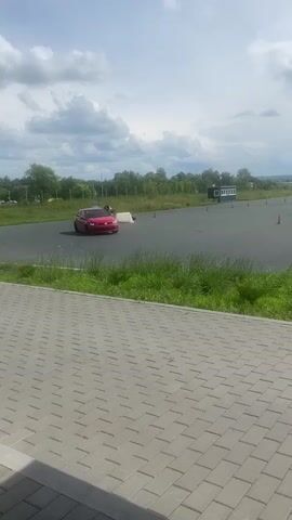 A red hatchback car is seen driving on an asphalt surface, possibly a racetrack or driving course. The car is executing a maneuver, with its wheels turned. There are orange traffic cones placed on the asphalt, indicating a course or obstacle. In the background, there is green grass, trees, and a cloudy sky. A building and a large white tent-like structure are also visible in the distance. The foreground shows a tiled pedestrian area. The video appears to be a recording from a stationary viewpoint.