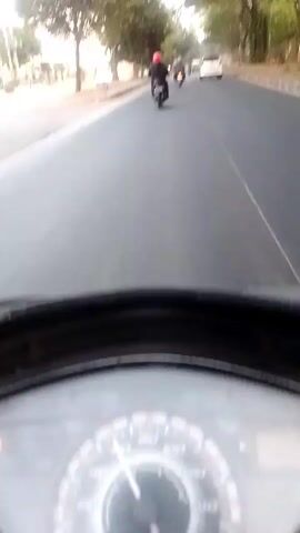 The video shows a first-person perspective of someone riding a scooter on a paved road. The rider is following a group of motorcycles. The motorcycles are in a line, and the lead motorcycle has a rider wearing a red helmet. There are trees lining the side of the road, and some buildings or structures can be seen in the background. The sky appears to be overcast or hazy. The scooter has a dashboard with a speedometer visible. The overall scene suggests movement along a road in an urban or suburban environment during daylight hours.