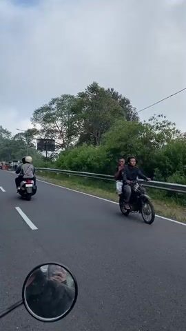 The video shows a road with a cloudy sky. There are trees and vegetation on the right side of the road. A motorcycle with two people on it is traveling in the same direction as the camera. A car is also visible in the distance on the left side of the road. There are several stalls or small shops lining the right side of the road. The camera appears to be mounted on another motorcycle, as a rearview mirror is visible in the foreground. The scene suggests a scenic route, possibly in a rural or mountainous area.