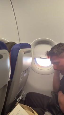 The video shows the interior of an airplane cabin, focusing on a window view. The window shade is partially open, allowing sunlight to stream in. Outside, there appears to be a view of clouds or land, possibly during sunrise or sunset due to the warm light. In the foreground, we see the side of an airplane seat with a blue headrest. The person seated next to the window can be seen looking outside.