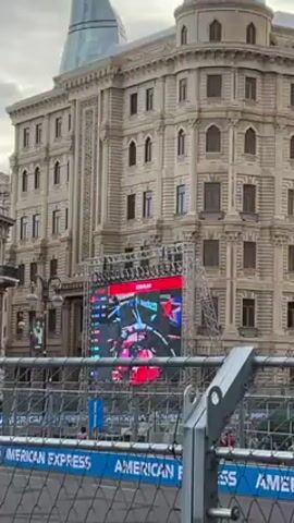 A large screen shows a Formula 1 race car driving the track during the Azerbaijan Grand Prix. The video is filmed from behind a safety fence marked with "AMERICAN EXPRESS". Behind the screen, there is a tall, ornate, tan building, likely in Baku, Azerbaijan. The sky is overcast.
