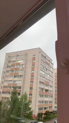 The video shows a view from an open window looking out at a cloudy sky and apartment buildings. On the right side of the frame, a white window frame and handle are visible. The buildings are multi-story and appear to be in a residential area, with trees and a playground visible in the foreground. The weather appears overcast.
