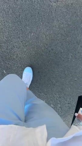 The video shows a person's legs and feet as they walk on an asphalt surface. The person is wearing light blue pants and white sneakers with light blue accents. A shadow of the person is cast on the asphalt, indicating it is daytime and sunny. The camera is angled downwards, focusing on the lower body of the person. The overall scene is outdoors on a paved path.