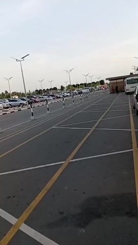 The video shows a parking lot filled with many cars on a sunny day. The parking spaces are marked with white lines, and yellow lines stripe the road. Black and white striped poles line the parking spaces. Tall light poles stand throughout the lot. A sign in a parking island warns, "NO PARKING UNAUTHORIZED VEHICLES WILL BE TOWED AT OWNER'S EXPENSE." Palm trees are visible in the background.