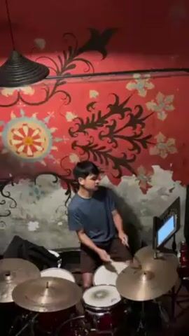 A young man is energetically playing drums in a room with a vibrant, stylized wall mural of red, orange, blue, and green. He's wearing a blue t-shirt and brown shorts. A drum set surrounds him, with a tablet on a stand in front, presumably displaying sheet music. A unique black hanging lamp adds to the room's character.