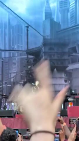 The video shows a live concert performance. A male singer, dressed in a military-style jacket with gold trim and black pants, stands center stage, holding a microphone. Behind him, a drummer plays a drum kit with a band logo visible on the bass drum. The stage features elaborate set design depicting a futuristic cityscape with tall buildings and metallic structures. A crowd of concert-goers can be seen in the foreground, many holding up their cell phones to record the performance. The lighting is dramatic, with a blue haze and spotlights.