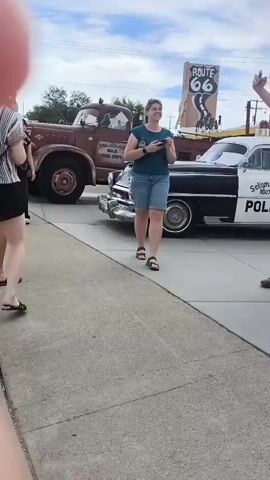 The video shows a man and a woman standing next to a vintage red truck with cartoon eyes painted on the hood. The truck is parked on a street, with a building labeled "Highway Hot Rods" and a Route 66 sign in the background. A classic black and white police car is parked behind the truck. The man is wearing a black t-shirt and blue shorts, and the woman is wearing a teal t-shirt and denim shorts. The man appears to be gesturing towards the truck, while the woman is looking at her phone. The overall environment suggests a tourist attraction or a car show related to Route 66.