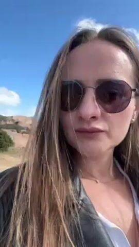 A woman with long brown hair, wearing dark sunglasses, a white shirt, and a black leather jacket, is looking at the camera. She is outside on a sunny day with a blue sky in the background. There are rock formations and a building visible in the distance. The wind is blowing her hair. She smiles slightly.