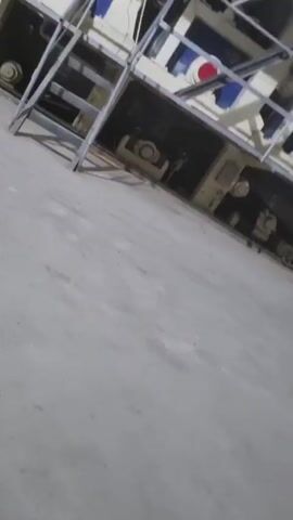 The video shows a view from a high vantage point within a large industrial facility. The camera initially focuses on a section of the building's machinery, featuring pipes, tanks, and structural metalwork. The camera then pans upward and outward, revealing a wider view of the facility. In the distance, a forklift truck is visible. The space has a high ceiling with exposed metal beams. Large containers are stacked in the foreground, below the camera. Overhead, a red industrial crane spans the width of the building. The camera then shifts, offering a closer perspective of a vertical wall beside the initial machinery setup.