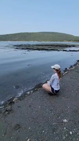 A young woman sits cross-legged on a pebbly beach, facing a calm body of water. She wears a white cap, a long-sleeved rash guard, and dark shorts. Her right hand is extended towards the water. Several concentric ripples are visible on the surface of the water in front of her, indicating something has just been thrown into it. The water is dark and reflects the sky. In the background, a tree-covered hill slopes down to the water's edge. The sky is a pale blue, with no clouds visible.