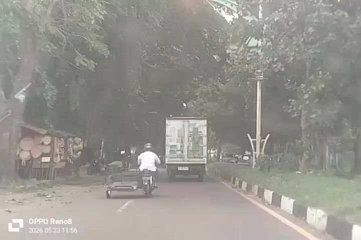 The video shows a road with trees lining the sides, a white truck, and a motorcycle with a sidecar, traveling in the same direction. There are parked cars and other motorcycles on the side of the road. A person walks on the side of the road. The video is shot from a vehicle following the motorcycle and truck. The date and time stamp at the bottom of the video reads "OPPO Reno8 2026.05.23 11:56".