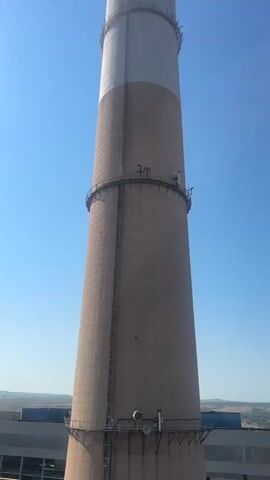The video shows a tall, cylindrical smokestack against a clear blue sky. The smokestack is constructed of concrete segments and has a light brown color for the majority of its height, transitioning to a lighter cream or white color towards the top. Two distinct bands are visible encircling the smokestack. One band is located near the bottom, and another band is situated higher up, just below the color transition. A vertical ladder is attached to the side of the smokestack, running from near the bottom to one of the bands. Small platforms or maintenance access points are visible at various levels on the smokestack, particularly around the bands. The base of the smokestack appears to be supported by a wider, concrete structure, parts of which are visible at the bottom of the frame. The surrounding environment is not clearly visible but appears to be flat terrain with some distant structures. The camera angle is looking upwards at the smokestack, emphasizing its height and scale. The lighting suggests a sunny day.