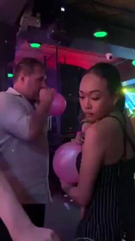 In a dimly lit environment, possibly a club, a man and a woman are seen holding pink balloons. The man, wearing a light-colored shirt and dark pants, is blowing into his balloon. The woman, in a pinstripe top and black straps, is holding hers. They're standing close to each other, and the scene seems to be part of a social gathering with music playing in the background. The club's setting includes speakers, red and pink lighting, and a stage visible in the background. The woman's hair is pulled back, and she has tattoos on her back.