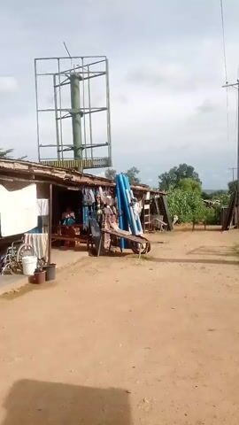 The video begins by showing a dusty, unpaved area next to a row of small shops. A large, empty billboard structure stands to the left, and a utility pole with wires is visible on the right. Blue pipes are leaning against a wooden structure in front of one of the shops. The camera then pans to the right, revealing a silver sedan parked on a dirt road. A stack of blue pipes is also visible on the side of the road. As the camera continues to move, it captures a green truck with a flatbed trailer driving down the road. Two people appear to be in the truck bed. The video ends with a close-up of the truck as it drives away. The environment appears to be a rural or semi-urban area with unpaved roads and modest buildings.