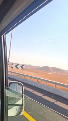 The video shows a view from inside a vehicle, looking out a side window. The vehicle appears to be traveling on a road next to a median barrier. The landscape outside consists of dry, golden grass, some scattered trees, and distant hills. There are power lines and utility poles visible against a clear blue sky. The sun is shining, casting shadows on the road. The time stamp indicates the video was recorded on October 24, 2023.