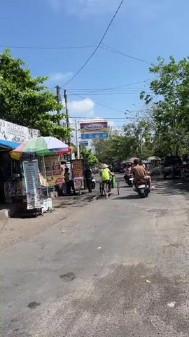 The video shows a street scene in Sepande, Indonesia, likely during the day. The weather appears to be sunny with a clear blue sky and some scattered clouds. The street is paved with asphalt, showing signs of wear and tear, with some cracks and rough patches.

Several people are visible. In the foreground, a person is riding a motorcycle away from the camera. Further down the street, another person is pedaling a bicycle with a passenger or cargo in a trailer attached to the back. There are also people standing near stalls or shops lining the side of the road.

The scene is bustling with activity. Along the left side of the street, there are numerous small stalls or shops selling various goods, some displaying colorful signs and advertisements. One stall has a prominent sign with text, and another appears to have a menu or list of items displayed. On the right side of the street, there are more vehicles, including a dark-colored SUV or van.

A prominent sign for "Indomaret" is visible in the distance, indicating the presence of a popular convenience store chain in Indonesia. This, along with the general streetscape and the presence of people and vehicles, suggests a commercial or residential area. Trees and foliage are scattered throughout the scene, particularly on the right side, adding to the tropical feel.

The overall atmosphere seems to be that of a lively, everyday street scene in a busy town or city. The lighting and clear sky suggest it is daytime, likely mid-morning or afternoon. There are no obvious signs of distress or unusual events; the mood appears to be casual and ordinary. The activity includes transportation, commerce, and people going about their daily routines.