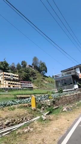 The video pans across a street scene with a clear blue sky overhead. On the left, there's a two-story building with a modern design, featuring a balcony, large windows, and what appears to be a restaurant or cafe. Below it, a stone wall borders a terraced field with green crops. Further up the hill, there are more buildings and trees. On the right side of the street, there is a road with a few vehicles, including a car and a truck, and more buildings, some with signs. Power lines are visible throughout the frame. The scene suggests a rural or semi-rural location, possibly a tourist area, with a mix of residential, commercial, and agricultural elements.