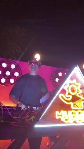 A man with sunglasses and headphones is standing behind a DJ setup, his hands hovering over the equipment. He is wearing a black t-shirt. To his right, a neon sign in the shape of a triangle with a yellow figure and the word "HYPE" written in a stylized font is visible. The background is dimly lit with orange and red lights, and some hexagonal light fixtures. The overall environment appears to be a nightclub or a performance venue.