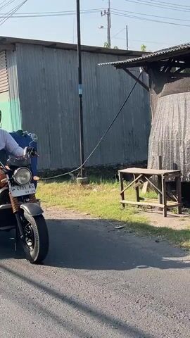 A motorcycle with a rider is turning right onto a dirt road. The rider is wearing a white shirt and black pants. The motorcycle is black and has a license plate that reads WM 6751 KE. In the background, there is a corrugated metal building and a wooden table. The scene is set during the daytime.