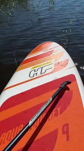 The video begins with a close-up view of a stand-up paddleboard on dark, rippling water. The paddleboard is white with orange and red stripes and features the "HYDRO-FORCE" logo. A paddle rests on the board. The camera then pans up and out, revealing a vast, calm lake under a bright blue sky dotted with fluffy white clouds. Reeds and tall grass grow along the shoreline in the foreground. In the distance, the opposite shore is lined with trees and a few buildings, suggesting a rural or semi-rural setting. The sun is shining brightly, creating shimmering reflections on the water's surface. The overall atmosphere is peaceful and serene, indicative of a pleasant day for outdoor recreation. The visible text on the paddleboard reads "HYDRO-FORCE" and "HT". The location is identified as Lendži, Latvia. The activity appears to be a peaceful paddle on a lake.