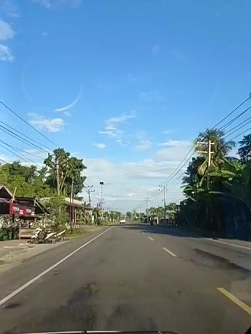 The video shows a forward-facing view from a vehicle traveling on a paved road. The sky is blue with scattered white clouds. On either side of the road are utility poles with many wires running between them. There are trees and buildings lining the road, suggesting a rural or suburban area. A motorcycle is visible on the left side of the road in the foreground. In the distance, other vehicles and more buildings can be seen. The road has yellow and white markings.