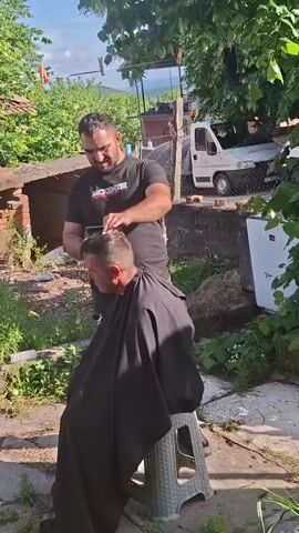 A man is giving another man a haircut outdoors. The barber stands behind the seated client, using clippers to trim his hair. The client is draped in a black cape. Both men appear to be middle-aged. The barber is wearing a black t-shirt with the word “MCKENZIE” printed on it. The client is sitting on a gray plastic stool. The setting appears to be a backyard, with green vegetation and a partial brick structure in the background. A white truck and a clothesline with laundry are visible in the distance. The weather appears to be sunny.