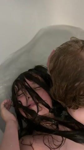 A close-up, overhead shot shows two young children in a bathtub. The child in the foreground, with long dark hair, is submerged in water up to their neck. Their eyes are closed, and their mouth is open, appearing to be gasping for air or experiencing distress. Their hand is raised, touching their face. Another child, with lighter brown hair, is positioned behind the first child, with their head close to the first child's face, obscuring some of the view. Water is splashing around the children. The bathtub appears to be white and filled with water.