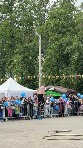 A bare-chested man with a large tattoo on his back, wearing black pants, is standing atop a tall unicycle in front of a crowd behind a metal barrier. The man is holding his arms out to his sides, then he falls forward and executes a front flip, landing on the ground on his feet. The crowd, separated by a barrier decorated with blue balloons, cheers and applauds. A referee in a black and green uniform with the number 15 watches closely. The event takes place outdoors in a paved area, bordered by green trees and decorations. A white tent is visible to the left, along with an inflatable brown sculpture in the background. A large metal ring lays on the ground near the unicycle.
