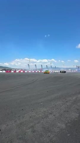 Two sports cars are drifting on a dirt track, emitting smoke. One car is orange and green, and the other is yellow and black. They are driving in opposite directions in a figure-eight pattern. Spectators are visible in the background, along with banners and flags. The sky is clear and blue.