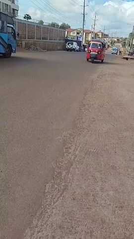 The video shows a red auto rickshaw driving towards the camera on a dirt road, then passing by. A blue bus or truck is parked on the side of the road. The rickshaw has the license plate KTW8382U. There are buildings and power lines visible in the background. The sky is partly cloudy.